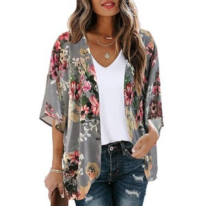A woman wearing a white V-neck t-shirt and a floral print kimono with varying shades of pink, green, and grey flowers. She also has on ripped blue jeans.