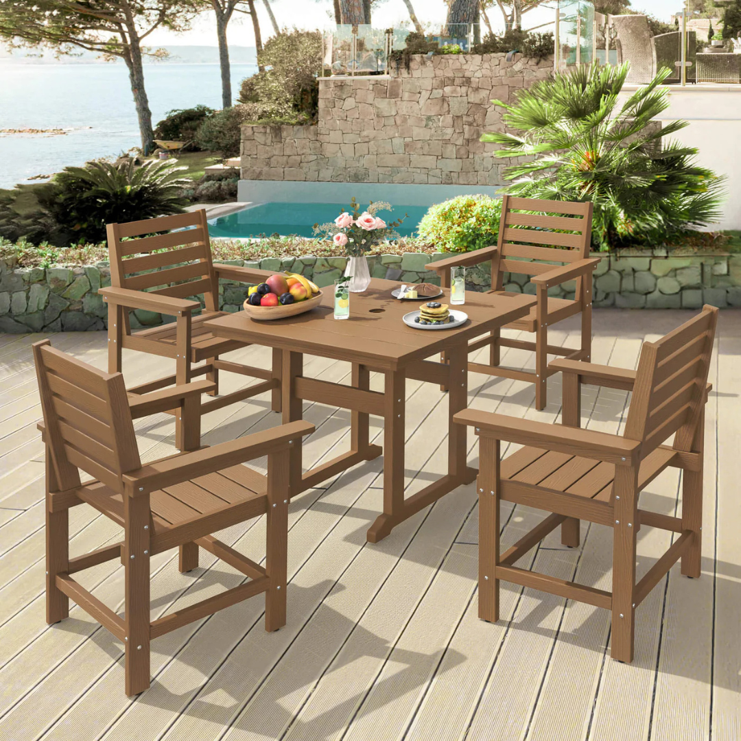 Patio dining set with a wooden table and four matching chairs on a deck, featuring a vase of flowers and plates with drinks. Background includes a pool and a stone wall with greenery.