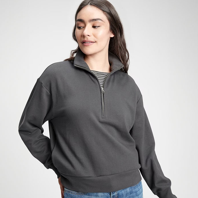 A woman models a dark gray half-zip sweatshirt with a high collar, ribbed striped inner detailing, and a relaxed fit. She pairs it with casual blue jeans.