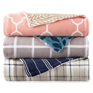 Three stacked blankets with different patterns: pink with white hexagons, gray with white squares, and blue with plaid. Each blanket shows a contrasting pattern on the fold.