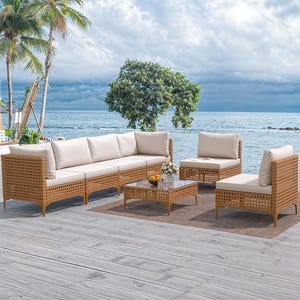 A patio conversation set featuring a three-piece sectional sofa, two armchairs, and two tables, all with tan wicker frames and white cushions, set on an outdoor deck by the sea.