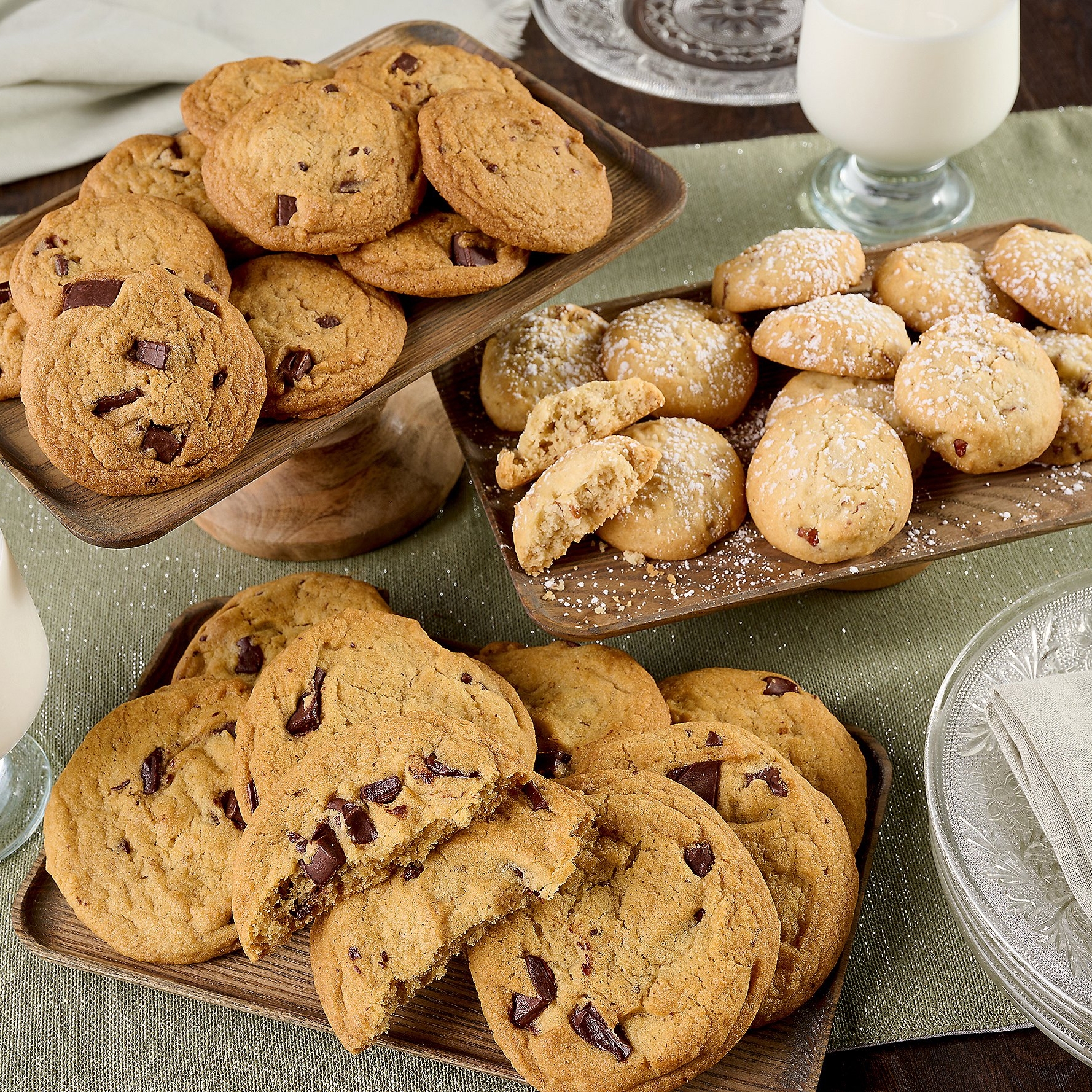 Assorted baked goods including chunky chocolate chip cookies and sugar-dusted round cookies, presented on wooden trays alongside glasses of milk.