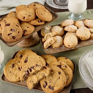 Assorted baked goods including chunky chocolate chip cookies and sugar-dusted round cookies, presented on wooden trays alongside glasses of milk.