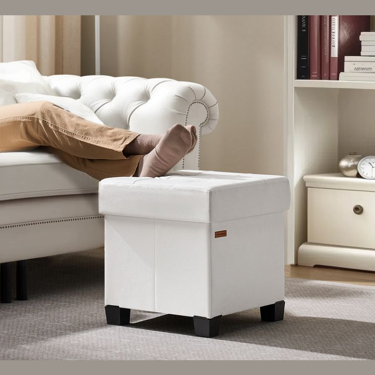 A white leather storage ottoman with black legs is shown in a living room setting, serving as a footrest. A person is lying on a white tufted sofa with their legs extended toward the ottoman.