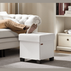 A white leather storage ottoman with black legs is shown in a living room setting, serving as a footrest. A person is lying on a white tufted sofa with their legs extended toward the ottoman.