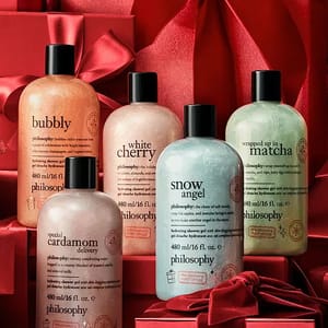 Bottles of Philosophy shower gel in various scents like Bubbly, White Cherry, Wrapped Up in Matcha, Special Cardamom Delivery, and Snow Angel are displayed among red gift boxes with ribbons.