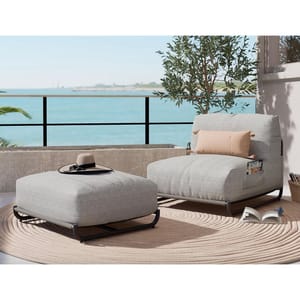 Outdoor lounge chair with matching ottoman, both in light gray fabric, on a circular beige rug, with coastal backdrop.