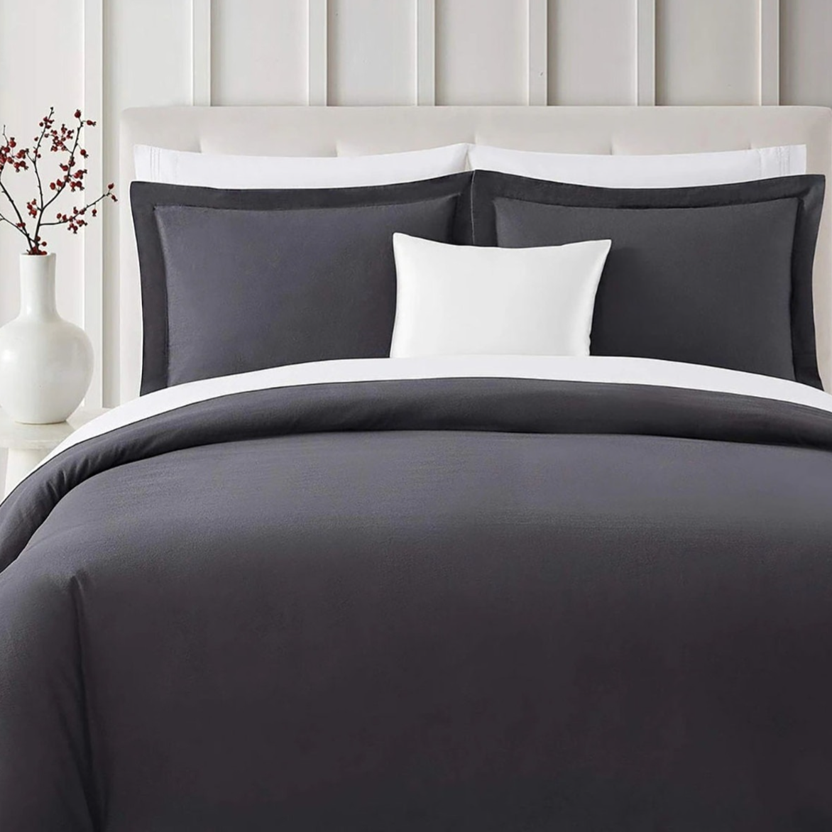 A neatly arranged bed features a charcoal gray comforter with white and gray pillows. A white vase with red berries sits on a bedside table against a white paneled wall.