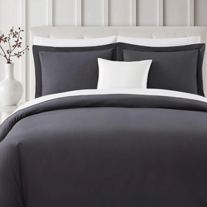 A neatly arranged bed features a charcoal gray comforter with white and gray pillows. A white vase with red berries sits on a bedside table against a white paneled wall.