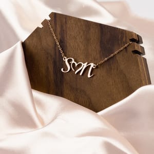 A rose gold necklace with the initials \“SM\“ hangs on a dark wooden holder against a white silk-like fabric backdrop.