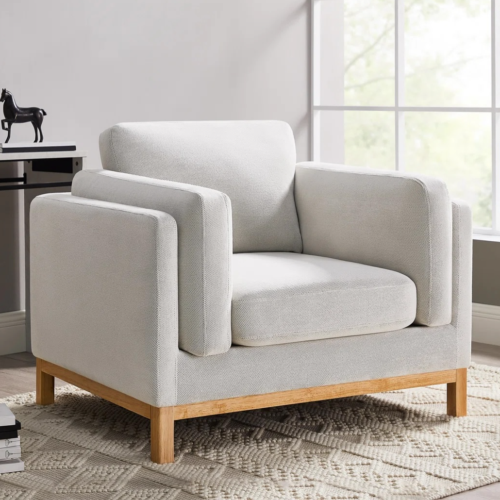 A modern armchair with a light grey fabric and wooden legs sits on a textured cream rug, near a window.