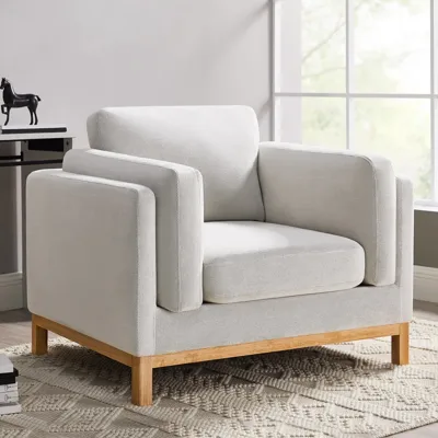 A modern armchair with a light grey fabric and wooden legs sits on a textured cream rug, near a window.