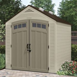 A beige storage shed with double doors featuring small windows on top and black handles. The roof is dark brown, and it's surrounded by plants and a paved area. Dimensions: 7 ft wide.