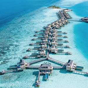 Overwater villas with private pools are connected by walkways over clear turquoise waters, leading to a small island with palm trees.