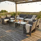 Outdoor patio set with rattan-style furniture, including a sofa, armchairs, and a glass-top coffee table, placed on a wooden deck under a pergola, decorated with string lights and throw blankets.