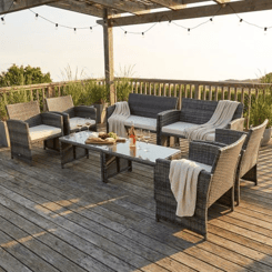 Outdoor patio set with rattan-style furniture, including a sofa, armchairs, and a glass-top coffee table, placed on a wooden deck under a pergola, decorated with string lights and throw blankets.