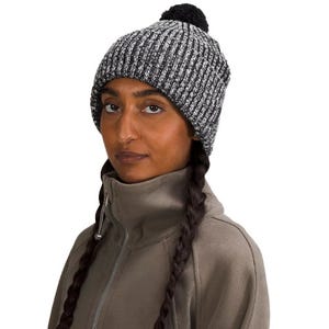 A woman is wearing a knit beanie with a pom-pom on top, featuring a fleece lining for warmth. The beanie is gray with black threading and a ribbed texture.