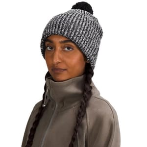 A woman is wearing a knit beanie with a pom-pom on top, featuring a fleece lining for warmth. The beanie is gray with black threading and a ribbed texture.