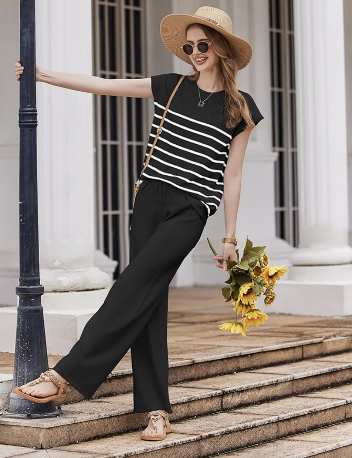 A woman in a black and white striped top, black wide-leg pants, and sandals holds sunflowers. Accessories include a straw hat, sunglasses, a necklace, and a crossbody bag.