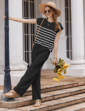 A woman in a black and white striped top, black wide-leg pants, and sandals holds sunflowers. Accessories include a straw hat, sunglasses, a necklace, and a crossbody bag.