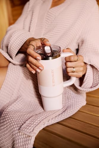 A person in a waffle-pattern robe holds a large white tumbler with a built-in straw and handle, featuring the brand \“Stanley.\“