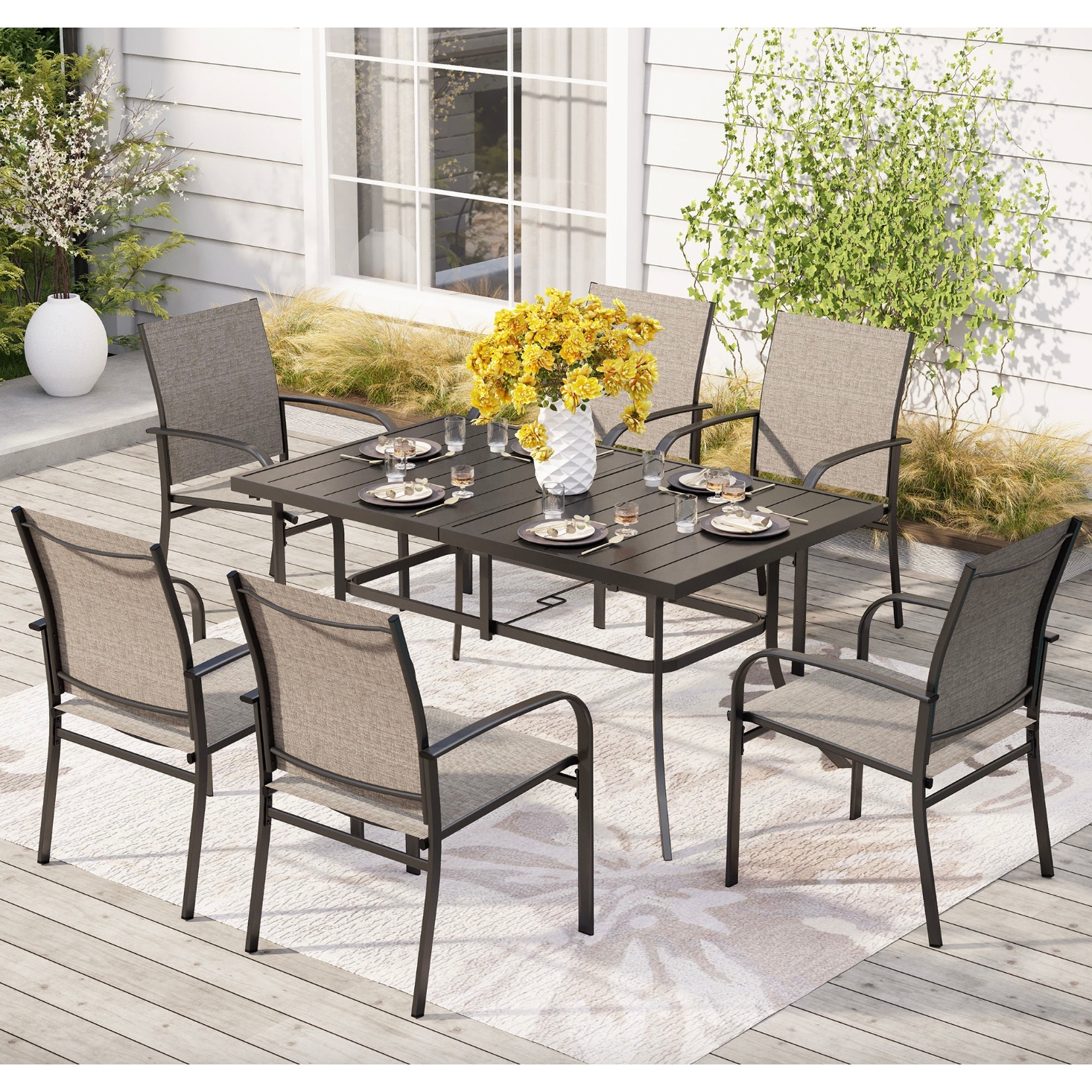 A 7-piece patio dining set includes a rectangular black metal table and six beige sling-back chairs. The table is set with plates and glasses, and a white vase with yellow flowers sits in the center on a light-toned outdoor rug.