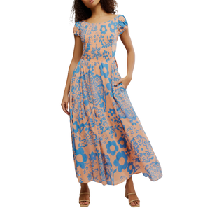 A woman is wearing a peach-colored maxi dress with bold blue floral patterns. The dress features short sleeves and a flowing skirt.