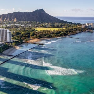 Coastal view with turquoise waters and waves, a sandy beach, lush greenery, and a prominent mountain in the background under a clear sky.