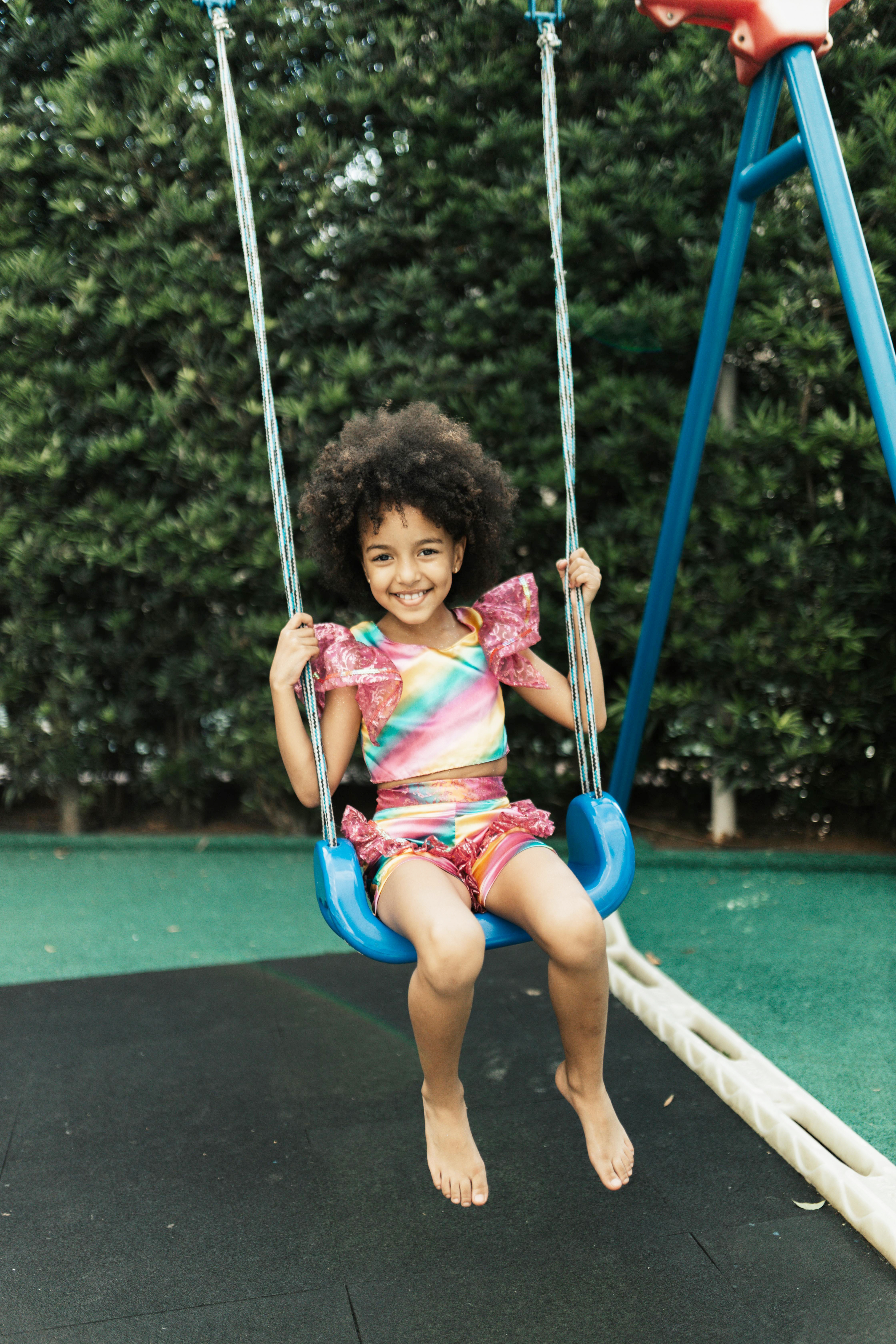 A smiling child on a blue swing set with a green protective floor and hedge in the background.