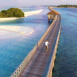 A long wooden boardwalk over clear turquoise water leads to a beach hut and lush island, with a person riding a bicycle along it.