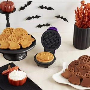 Halloween-themed mini waffle makers alongside ghost-shaped pancakes and waffles on a decorated table.