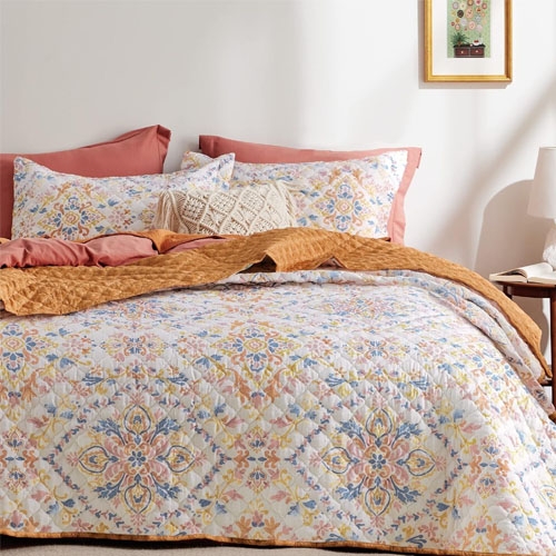 A colorful floral quilt on a bed with coordinating pillows and pillowcases, set against a neutral wall with framed artwork.