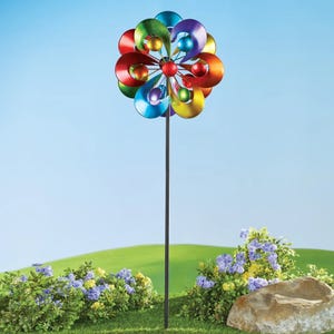 A colorful metal garden wind spinner with dual rotors on a stake set against greenery and blue sky.