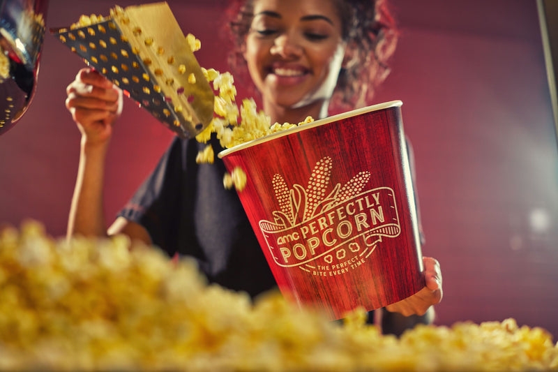 A person is filling a large AMC branded popcorn bucket with freshly popped popcorn.