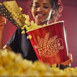 A person is filling a large AMC branded popcorn bucket with freshly popped popcorn.