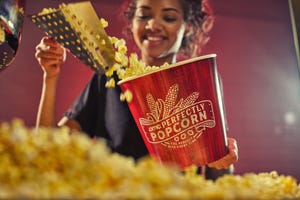 A person is filling a large AMC branded popcorn bucket with freshly popped popcorn.