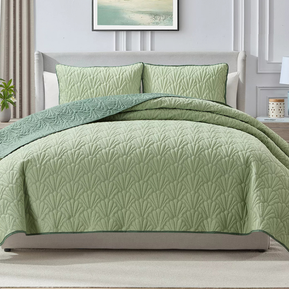 A light green quilted bedspread with a leaf pattern is displayed on a neatly made bed, accompanied by matching pillow shams. The bedding features a darker green trim and complements the light, neutral decor of the room.