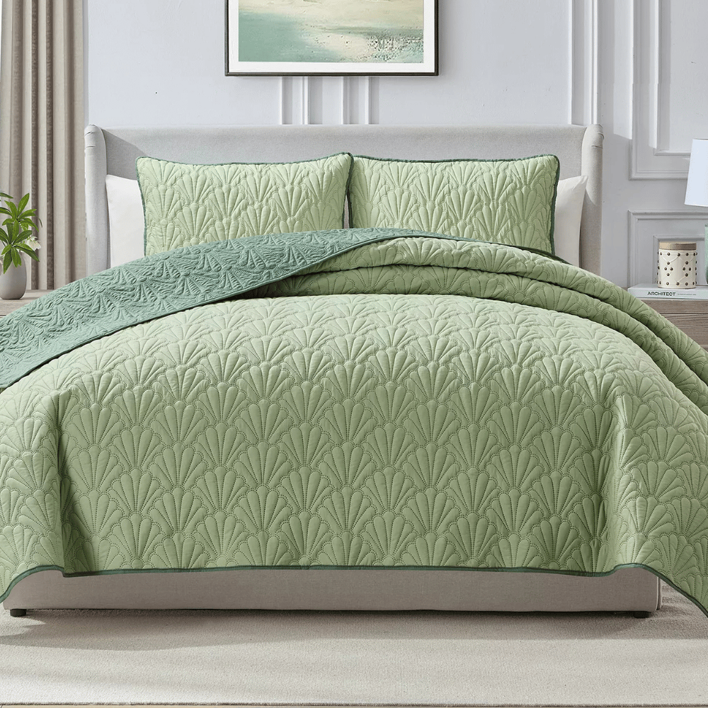 A light green quilted bedspread with a leaf pattern is displayed on a neatly made bed, accompanied by matching pillow shams. The bedding features a darker green trim and complements the light, neutral decor of the room.