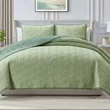 A light green quilted bedspread with a leaf pattern is displayed on a neatly made bed, accompanied by matching pillow shams. The bedding features a darker green trim and complements the light, neutral decor of the room.
