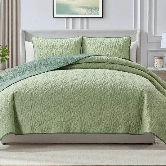 A light green quilted bedspread with a leaf pattern is displayed on a neatly made bed, accompanied by matching pillow shams. The bedding features a darker green trim and complements the light, neutral decor of the room.