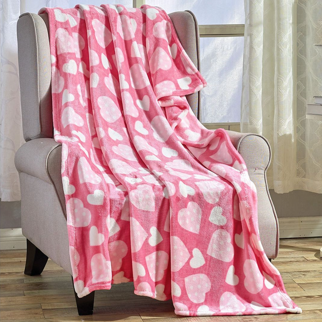 A pink fleece blanket with white heart patterns is draped over a light gray armchair, situated in a room with wooden flooring and large windows.