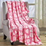 A pink fleece blanket with white heart patterns is draped over a light gray armchair, situated in a room with wooden flooring and large windows.