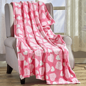 A pink fleece blanket with white heart patterns is draped over a light gray armchair, situated in a room with wooden flooring and large windows.