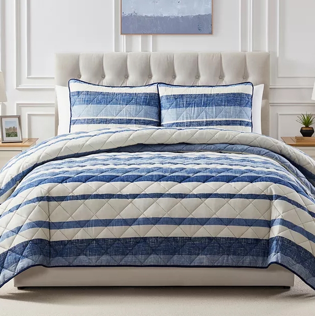 A neatly made bed features a quilted comforter with blue and white striped pattern and matching pillow shams. The headboard is upholstered and tufted, set against a paneled wall with minimalist decor.