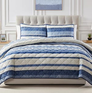 A neatly made bed features a quilted comforter with blue and white striped pattern and matching pillow shams. The headboard is upholstered and tufted, set against a paneled wall with minimalist decor.