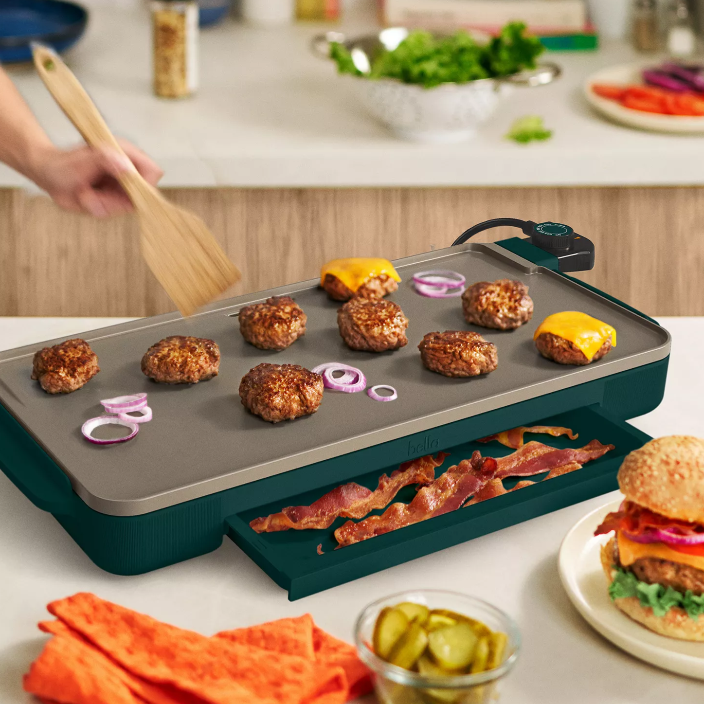 Electric griddle cooking burgers and onions, with bacon stored in a tray underneath. A completed cheeseburger, pickles, and condiments are nearby, set in a kitchen setting.