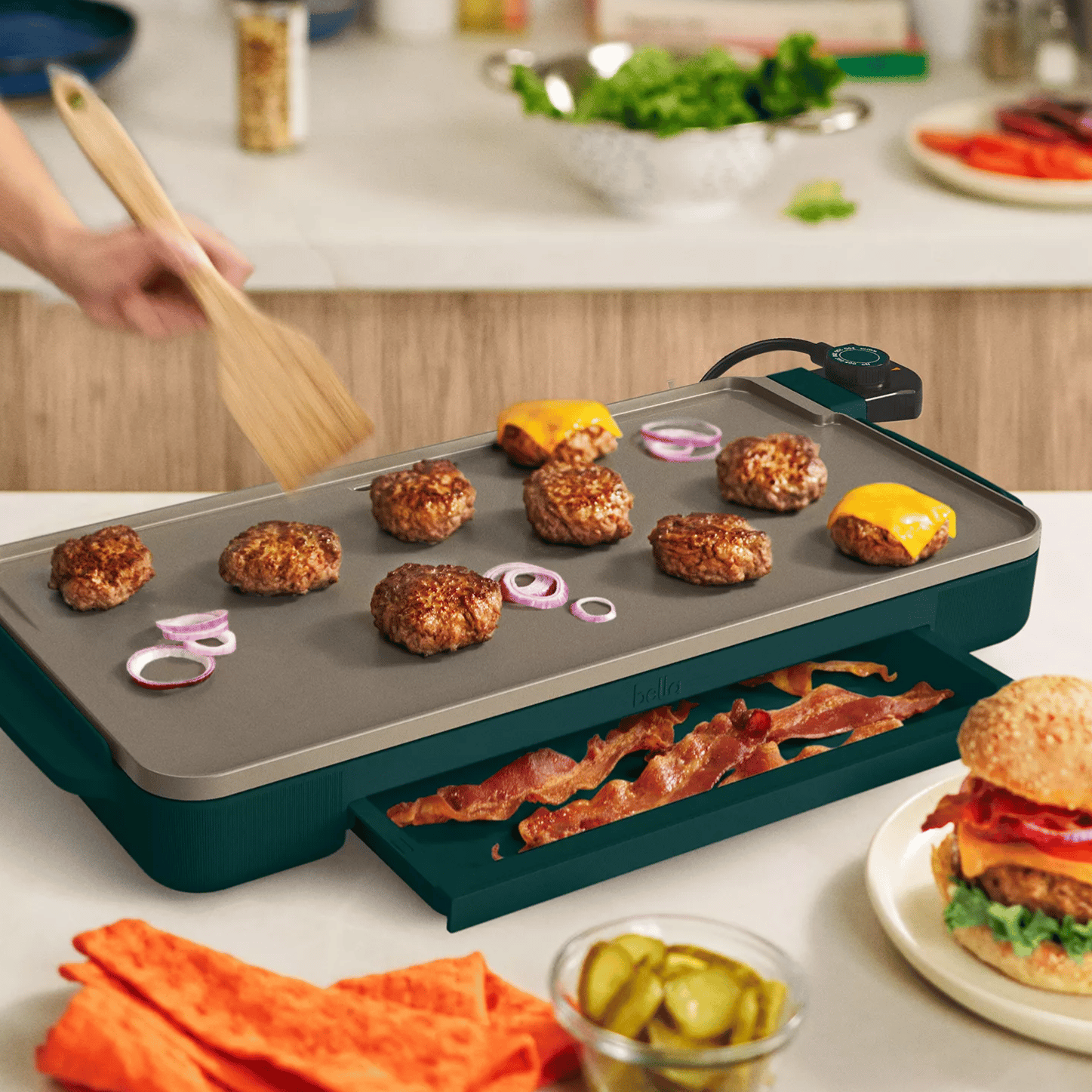 Electric griddle cooking burgers and onions, with bacon stored in a tray underneath. A completed cheeseburger, pickles, and condiments are nearby, set in a kitchen setting.