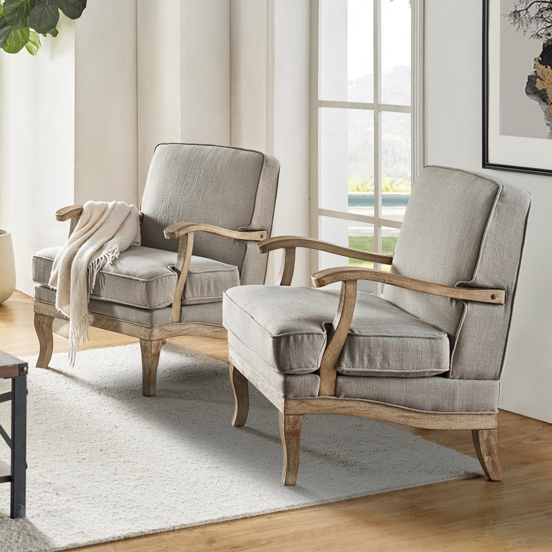 Two upholstered armchairs with light wood frames and neutral fabric cushions are positioned in a bright room with a large window, set on a textured rug near a plant.