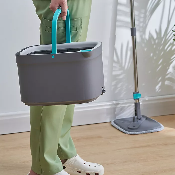 A person is holding a grey portable cleaning bucket with a blue handle, next to a flat mop with a cloth pad on a wooden floor.