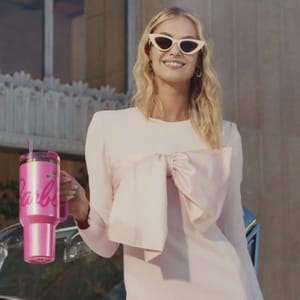 A woman is holding a pink Stanley tumbler with the Barbie logo; the tumbler has a lid and straw, designed for on-the-go drinks.